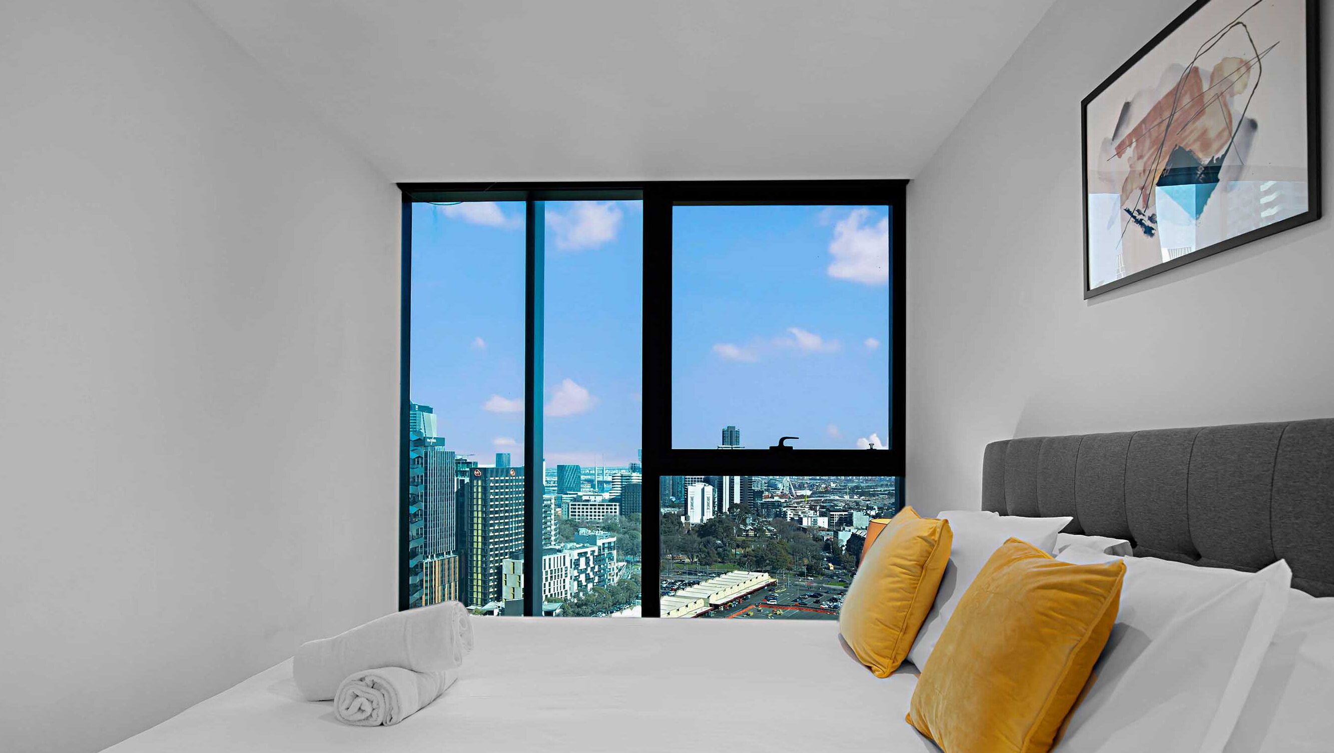 Habitación de hotel moderna con un amplio ventanal que ofrece vistas a la ciudad, una cama bien hecha con ropa blanca y un cabecero gris.