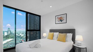 Ein Hotelzimmer mit einem großen Bett, zwei Nachttischlampen, einem gerahmten Kunstwerk und Blick auf die Stadt durch raumhohe Fenster.
