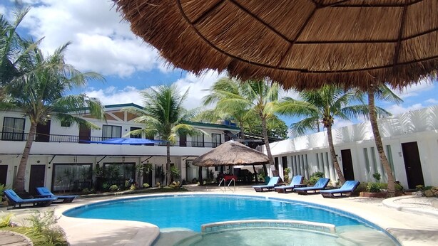 A pool area with a thatched umbrella, lounge chairs, and a building with balconies. - Casa Marta Hotel (Iloilo)