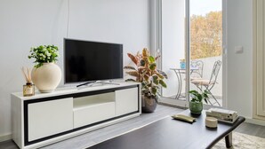 A modern living room with a flat-screen TV on a white cabinet, a coffee table with books and a remote, and a large window with a view of outdoor furniture. - Ribas Machico, a Home in Madeira (Machico)