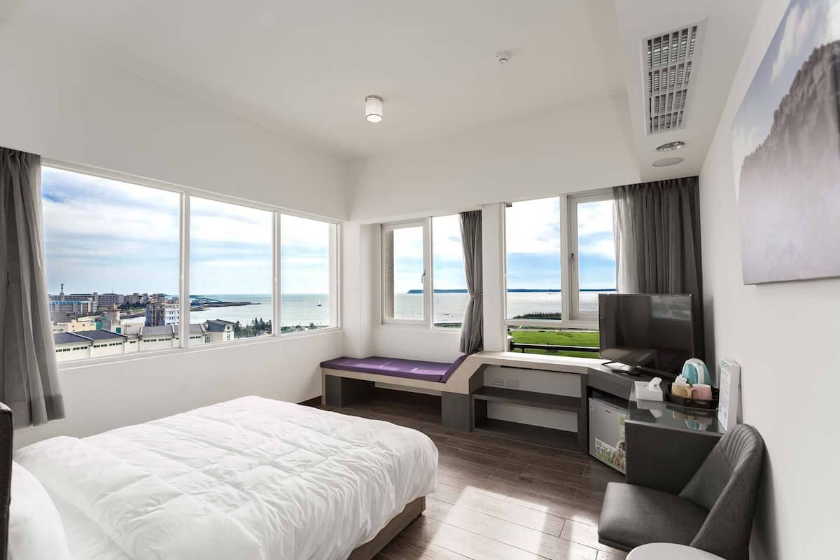 Ein Hotelzimmer mit einem großen Bett, einem Schreibtisch mit Stuhl, einem Fernseher und Blick auf Stadt und Wasser.