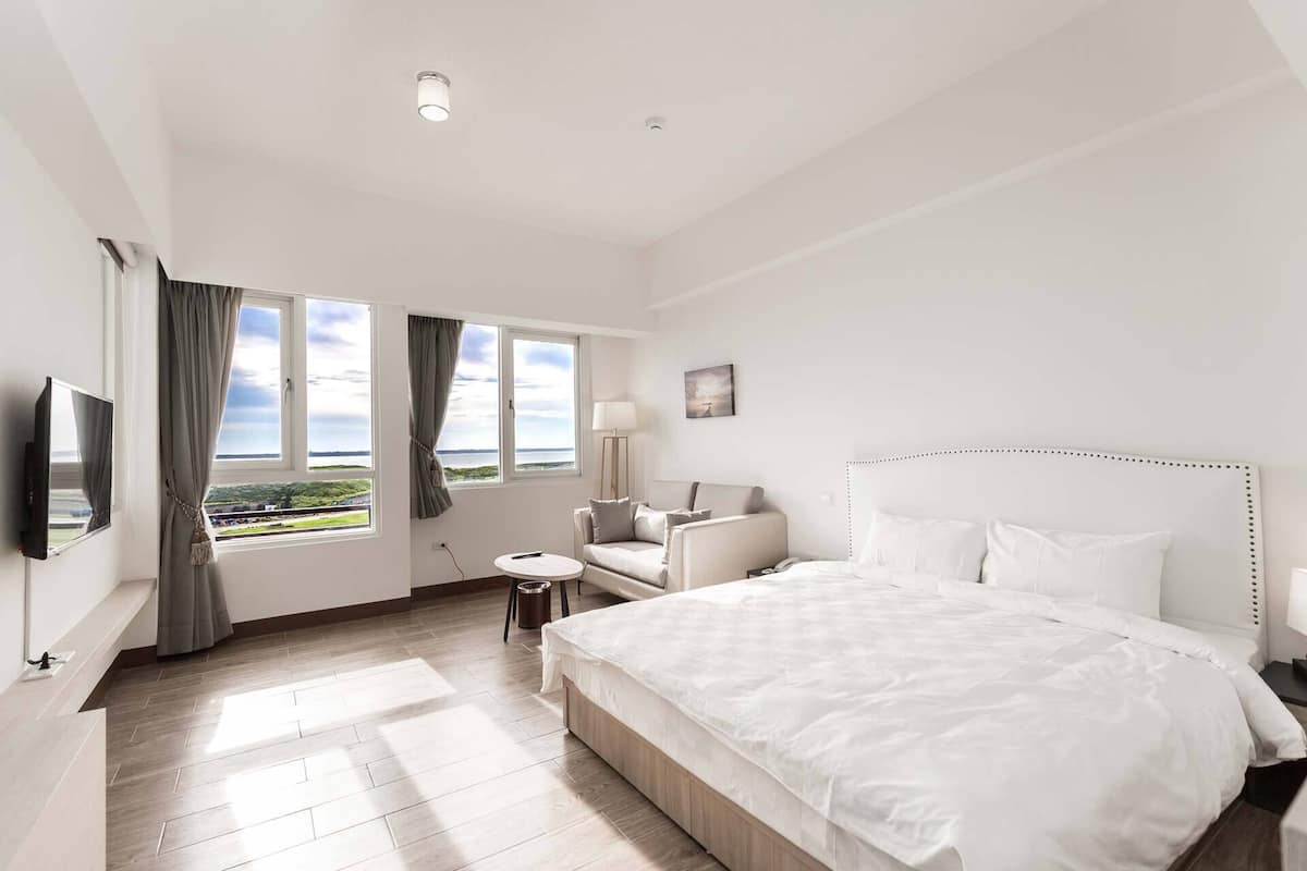 Ein Hotelzimmer mit einem großen Bett, einem Fernseher, einem Sessel, einem kleinen Tisch und Blick ins Freie.