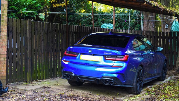 Een blauwe auto geparkeerd in een oprit met een houten hek en groen in de achtergrond.