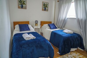 Ein Zimmer mit zwei Betten, jeweils mit blauer Bettdecke und weißen Kissen, einem kleinen Nachttisch mit Lampe sowie zwei an der Wand befestigten Bildern.