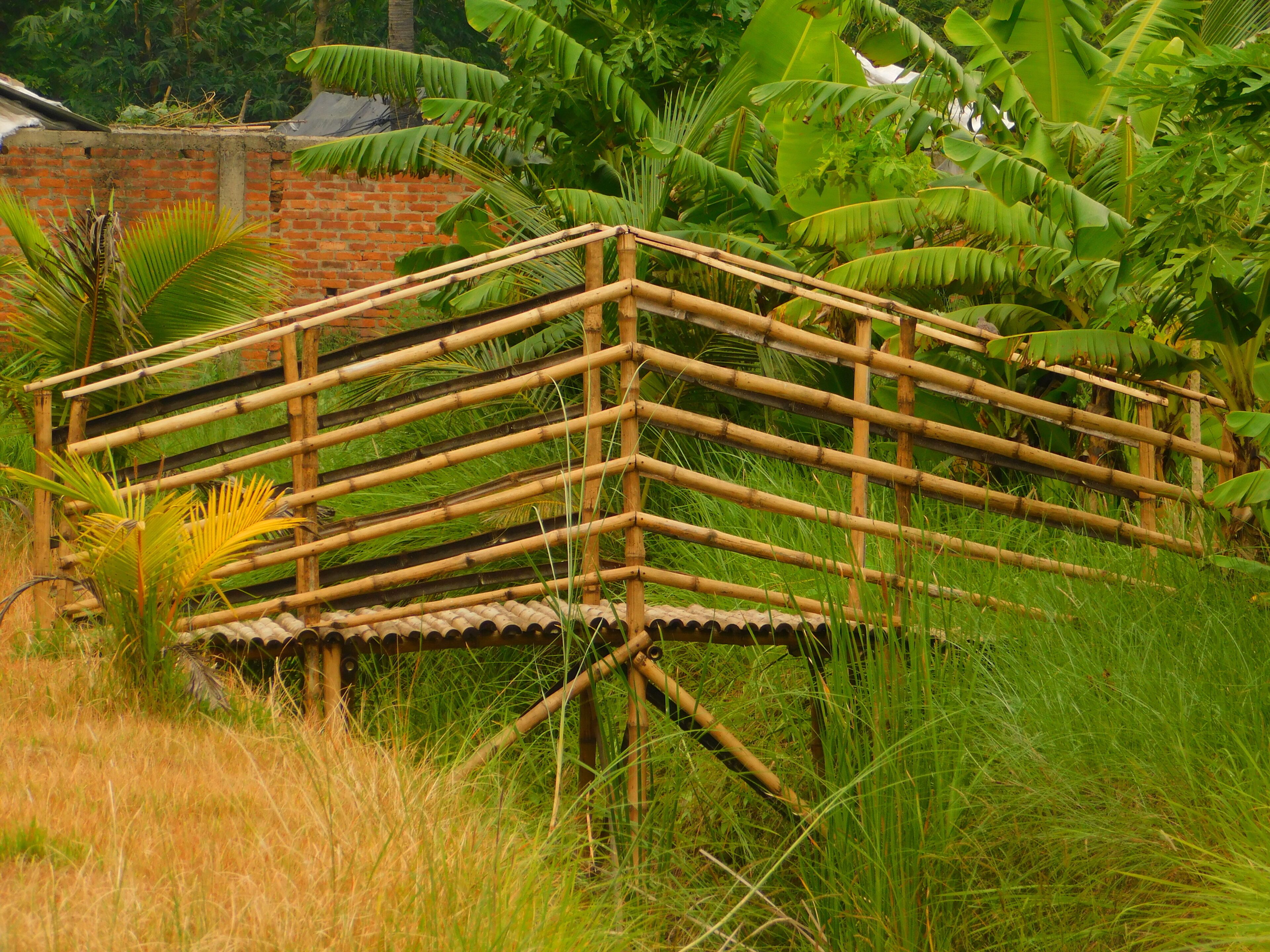 A bamboo structure in a lush garden with dense foliage and a brick wall in the background.