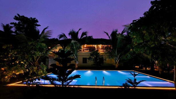A nighttime view of a hotel with a swimming pool, surrounded by palm trees and a lit building. - Gram Bangla Retreat (Alipur)
