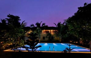 A nighttime view of a hotel with a swimming pool, surrounded by palm trees and a lit building. - Gram Bangla Retreat (Alipur)