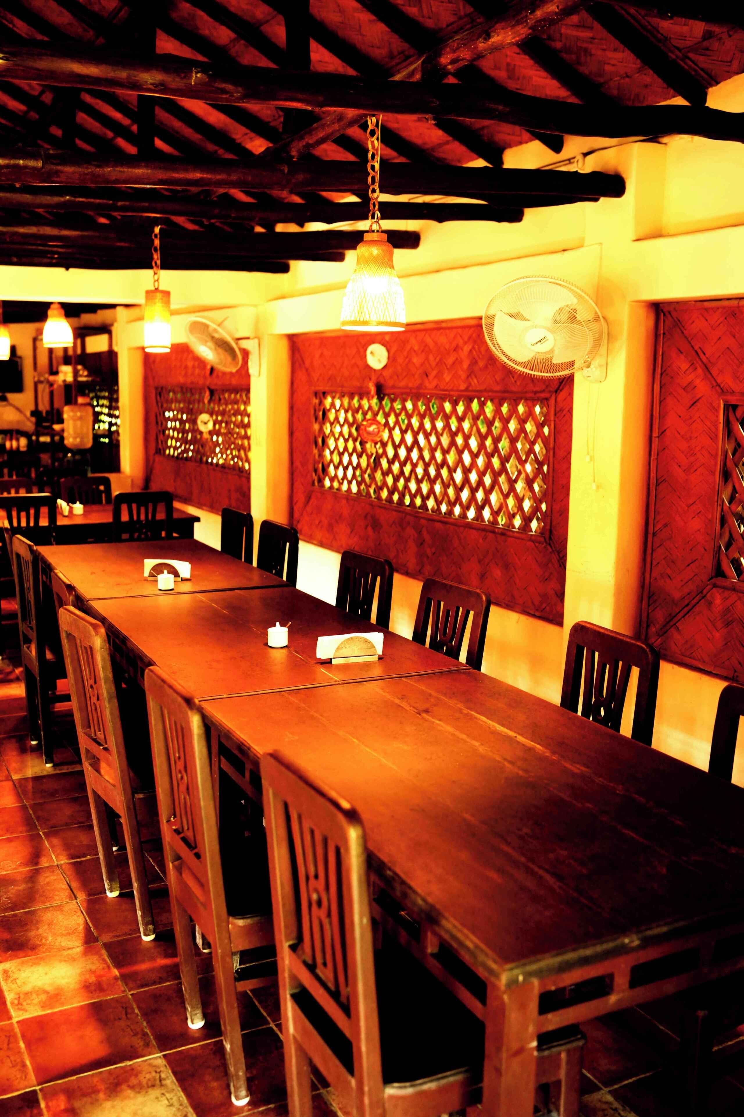 Una larga mesa de comedor de madera con sillas, un ventilador colgante y lámparas colgantes en un restaurante con vigas de madera y ventanas con celosías.