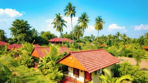 A tropical resort with multiple bungalow-style buildings, each featuring a red-tiled roof and surrounded by lush greenery and palm trees. - Gram Bangla Retreat (Alipur)