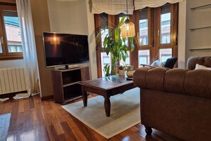 A living room with a TV, a brown sofa, a wooden coffee table, a potted plant, and large windows.