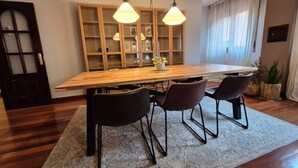 Uma sala de jantar com mesa de madeira, três cadeiras, iluminação pendente, uma estante e um vaso com planta.