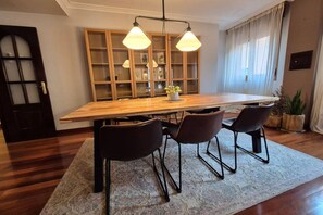 Zona pranzo con tavolo in legno, tre sedie, illuminazione a sospensione, libreria e una pianta in vaso.