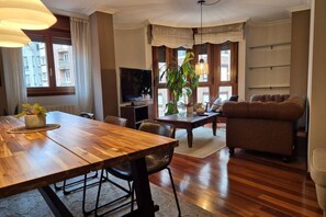 A living room with a wooden dining table, a sofa, a TV, and a large window with curtains. - 1A137 Apartamento con encanto puerto deportivo (Gijón)