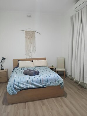 Un dormitorio con una cama, una mesita de noche, una silla, una decoración de macramé colgante y una ventana con cortinas.