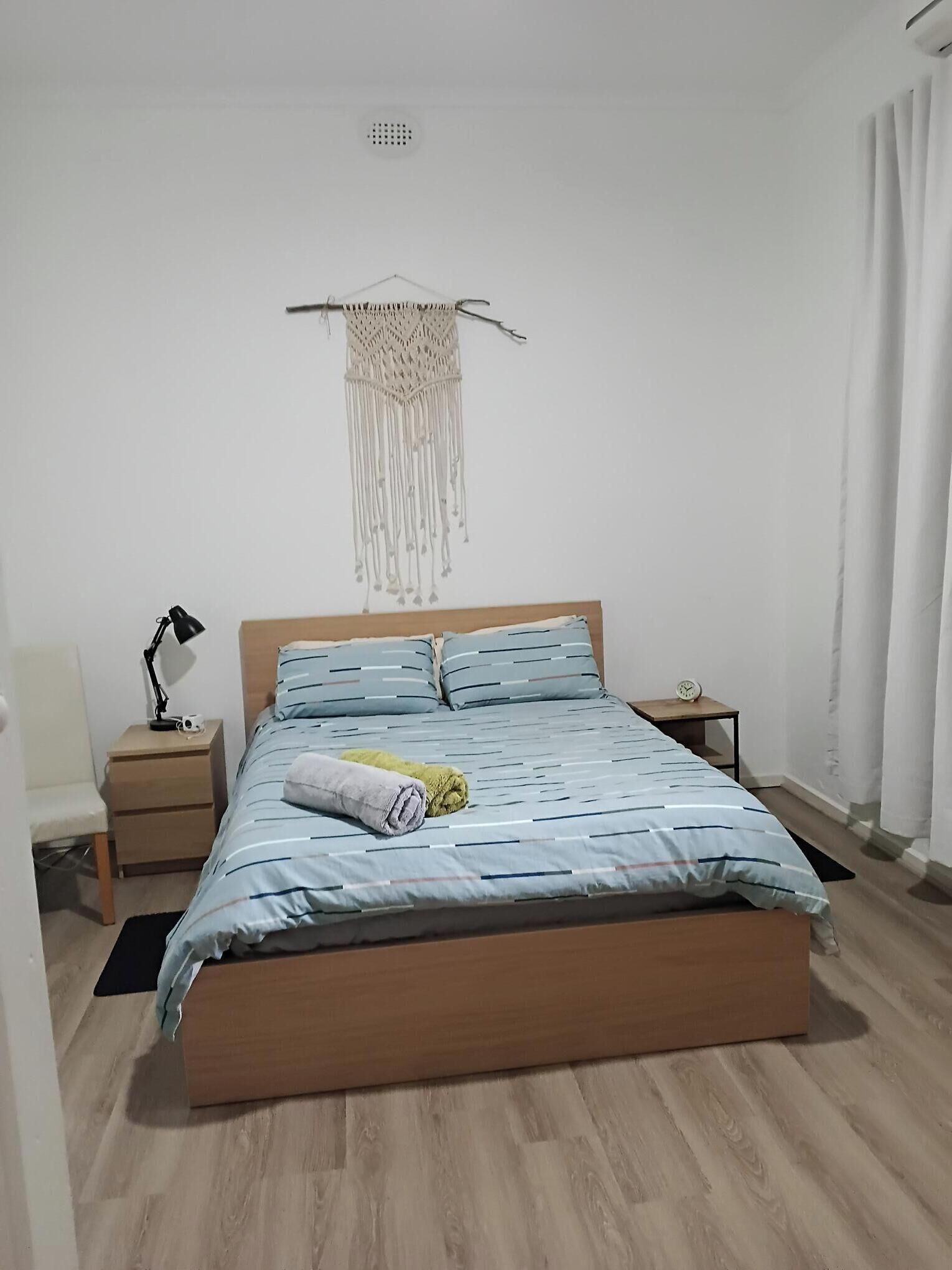 Une chambre avec un lit en bois, une table de chevet avec une lampe, une décoration murale en macramé et une petite horloge sur la table de chevet.