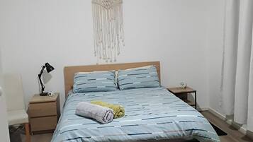 Une chambre avec un lit en bois, une table de chevet avec une lampe, une décoration murale en macramé et une petite horloge sur la table de chevet.