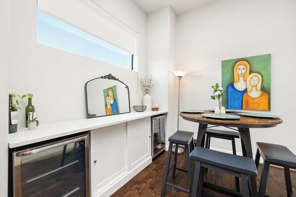 Une cuisine moderne avec des meubles blancs, un four encastré, un petit réfrigérateur et un coin repas comprenant une table ronde et des tabourets de bar.