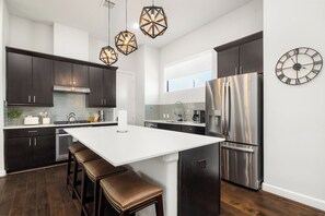 Une cuisine moderne avec des meubles de couleur sombre, un grand îlot central, des appareils électroménagers en acier inoxydable et des luminaires suspendus aux formes géométriques.