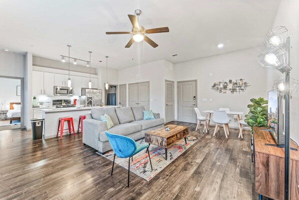A spacious living area with a grey sofa, blue chair, and red stools. A wooden coffee table is in the center. The room has a ceiling fan, pendant lights, and a modern kitchen in the background. - Cozy 2BR Suite Close to Downtown w Fast Wifi (Austin)
