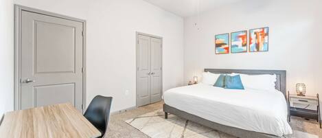 A bedroom with a bed, a desk, a chair, a ceiling fan, and artwork on the wall.