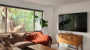 A modern living room with a large window, a sofa, a coffee table, and a TV on a wooden cabinet. - Family beach house among the gum trees (Ocean Grove)