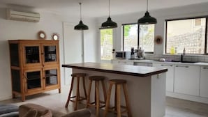 A modern kitchen with a central island, bar stools, pendant lights, and a wooden cabinet. - Family beach house among the gum trees (Ocean Grove)