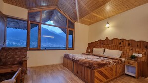 A bedroom with a wooden bed, a chair, a nightstand, and a large window with a view of mountains. - Redrosecottages  (Manali)