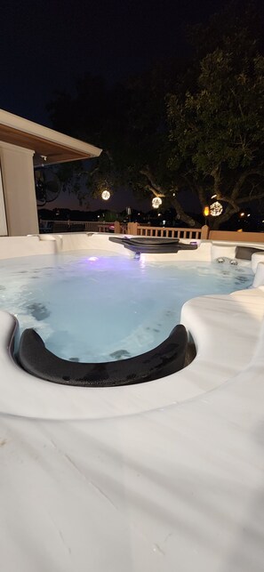 Een hottub met een gebogen rand en een klein watervalletje, omringd door witte ligstoelen.