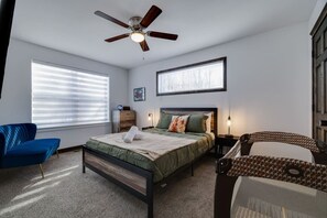 Quarto com cama, ventilador de teto, janela com persianas, uma cadeira, mesa de cabeceira e uma obra de arte na parede.