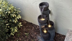 A tiered water feature with three illuminated tiers against a textured wall, surrounded by mulch and a small bush.