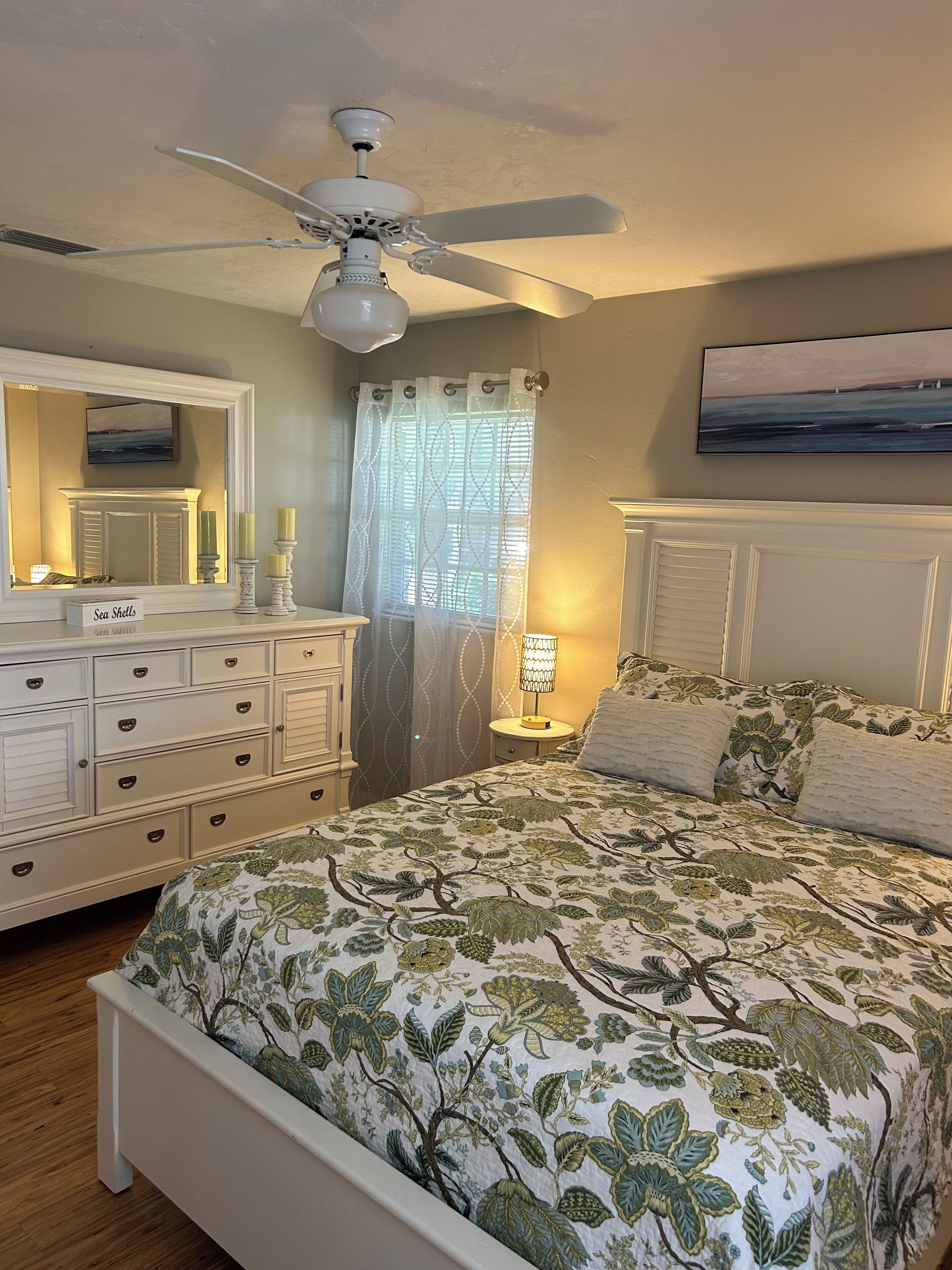 A bedroom with a bed, dresser, ceiling fan, and a window with curtains.
