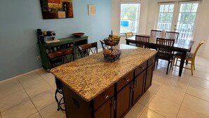 Una cocina con una isla central, un comedor con mesa y sillas, una pared azul con obras de arte y una ventana con persianas.