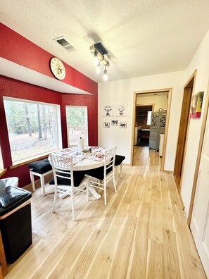 A dining area with a round table, white chairs, and a view of the outdoors through large windows.