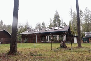 A wooden cabin with a sloped roof and skylights, surrounded by a fenced yard and tall trees.
