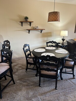 Un coin repas avec une table ronde, des chaises noires, un luminaire suspendu et une étagère murale agrémentée d’objets décoratifs.