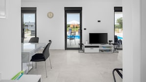 A modern living room with a flat-screen TV, a dining area with a glass table, and a view of a swimming pool and mountains.