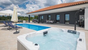A swimming pool with lounge chairs and a hot tub, a covered patio area, and a modern building with large windows.