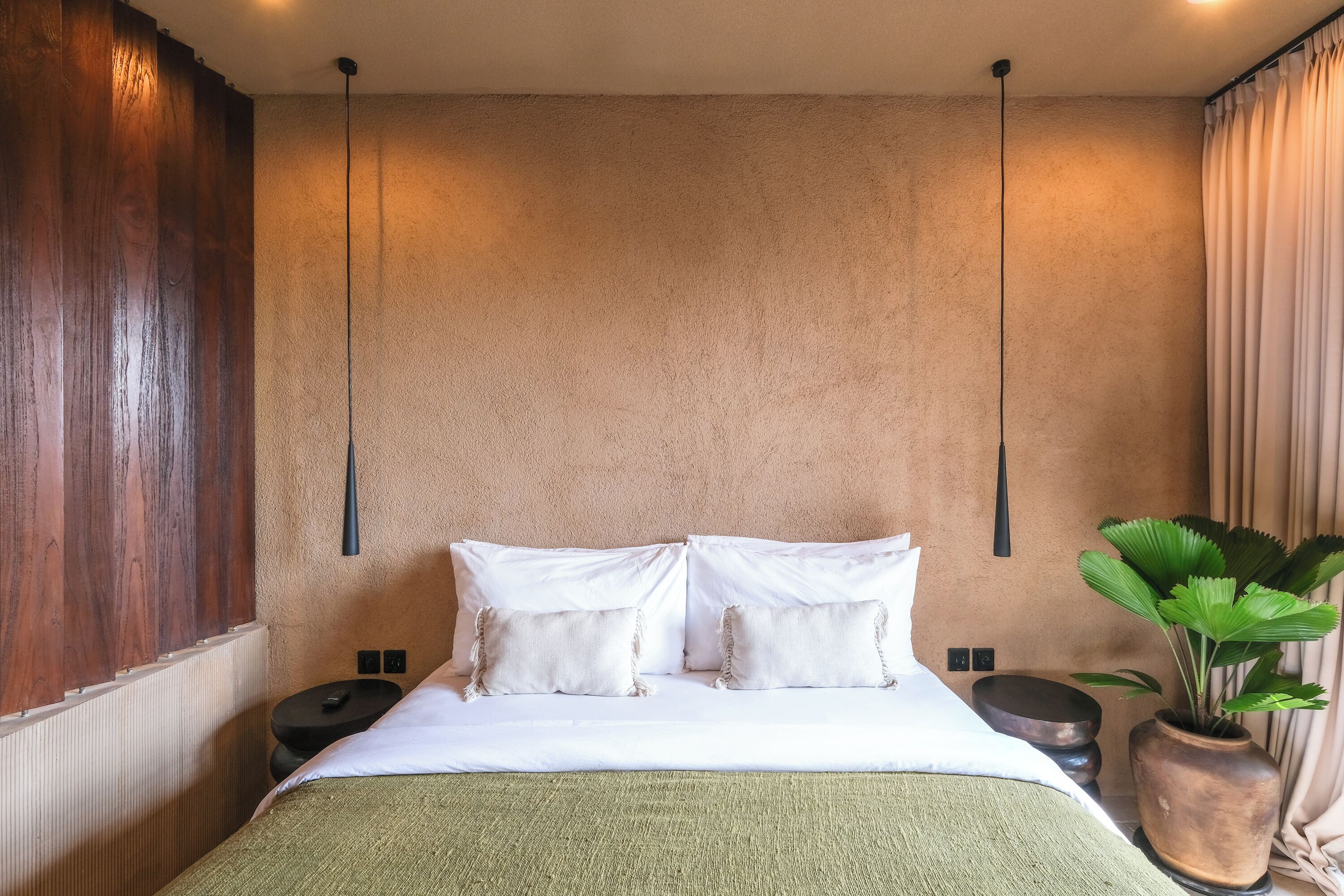 A neatly made bed with white linens and two pillows, a bedside table with a lamp, a potted plant, and a textured wall.