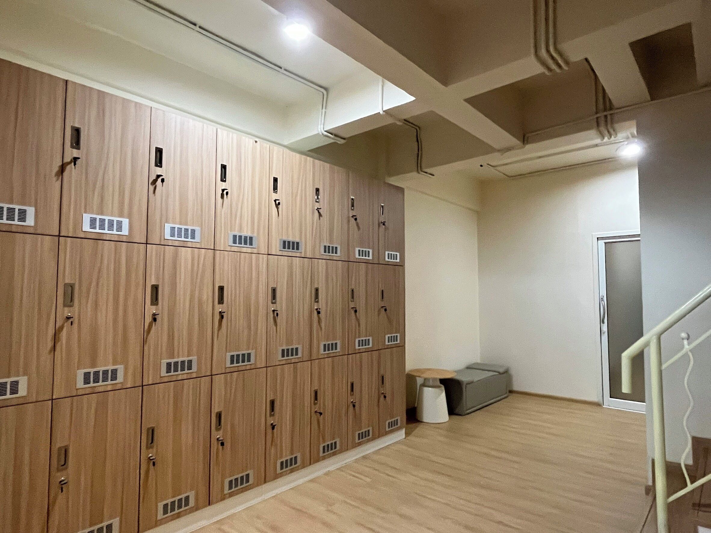 A row of wooden lockers with ventilation slits, a small trash bin, and a staircase with a metal railing.