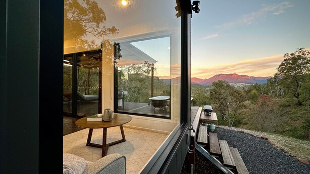 Ein modernes Interieur mit einem großen Glasfenster, einer Holzdielen Terrasse und Blick auf eine gebirgige Landschaft.
