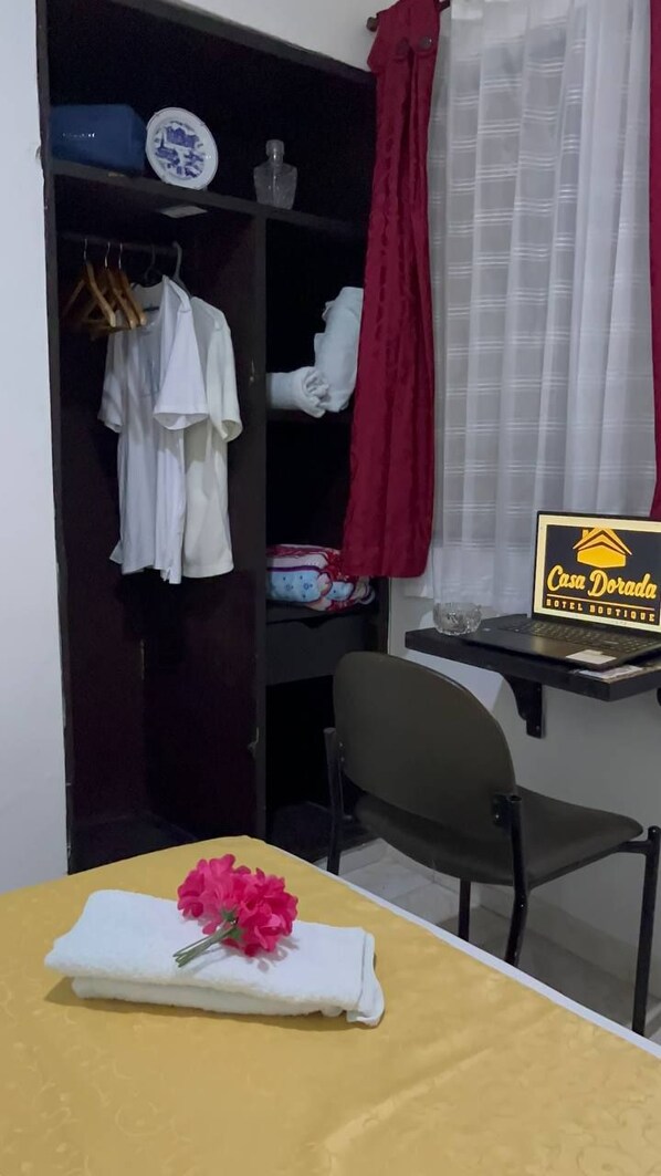 A hotel room with a bed, a table with a towel and flowers, a chair, a desk with a laptop, a wardrobe with clothes, a red curtain, and a window with white curtains. - Casa Dorada Hotel Boutique (Neiva)