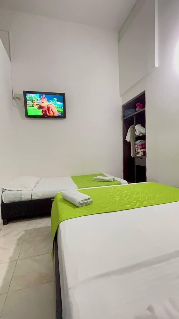 Une chambre d’hôtel avec deux lits, l’un recouvert d’une couvre-lit verte et l’autre d’une couvre-lit blanche. Un téléviseur fixé au mur diffuse un dessin animé.