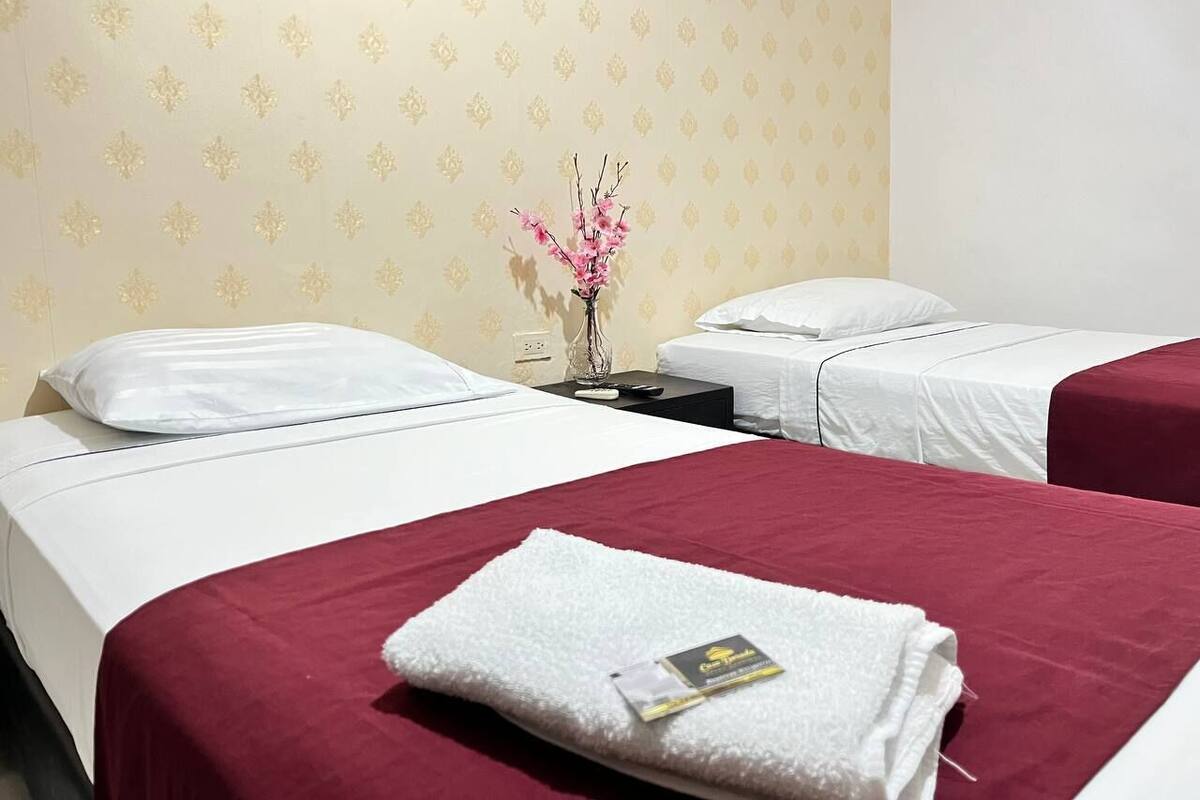 Une chambre d’hôtel avec deux lits, chacun doté d’un matelas blanc et d’une couverture rouge. Une petite table avec un vase rempli de fleurs roses se trouve entre les deux lits.