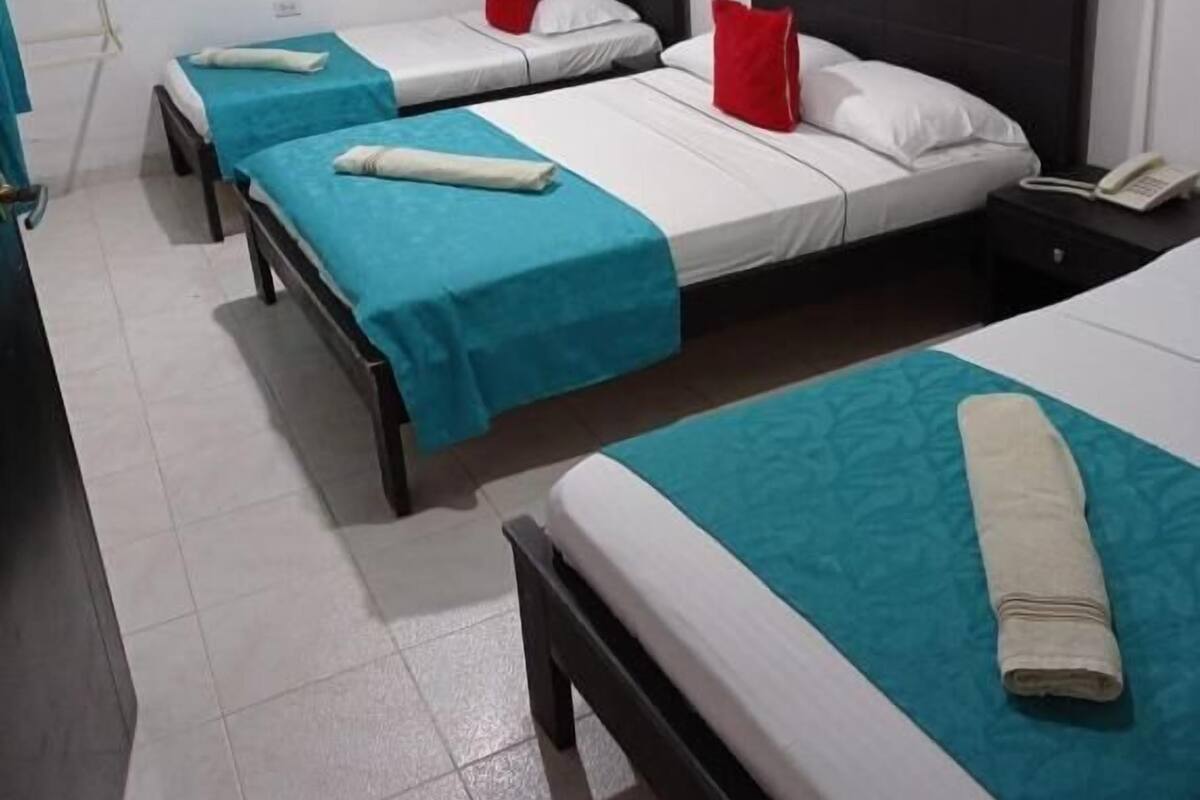Une chambre d’hôtel avec trois lits simples, chacun recouvert d’une couvre-lit turquoise et doté de draps blancs. Une table de chevet avec une lampe et un téléphone se trouve sur le côté droit de la pièce.