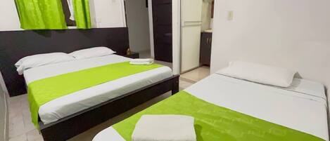Habitación de hotel con dos camas, cada una con una colcha verde y almohadas blancas. Hay una mesita con una toalla blanca encima. La habitación tiene una puerta que da a un baño y una kitchenette.