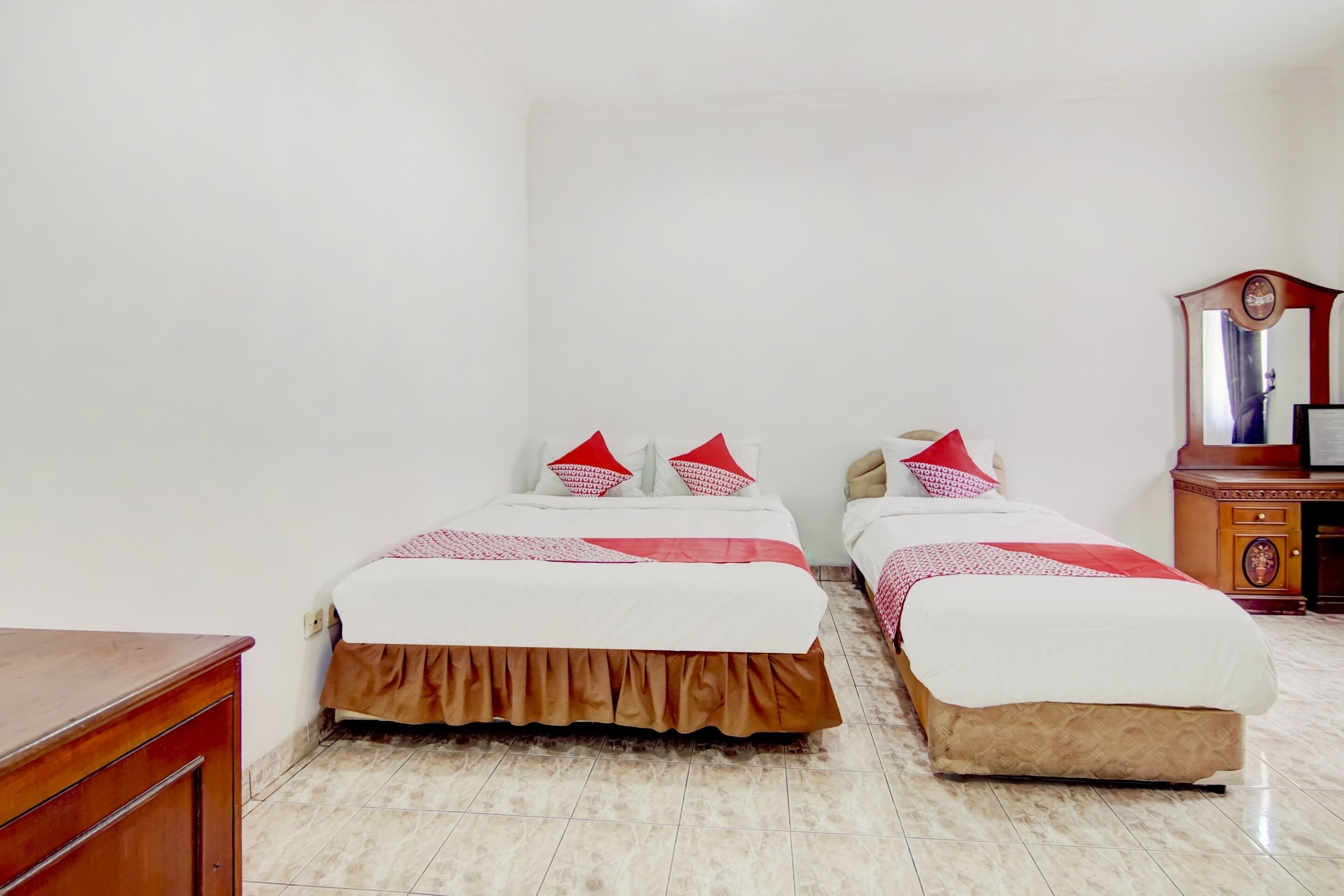 A room with two beds, each with a white bedspread and red pillows, a wooden dresser, and a mirror on the wall.