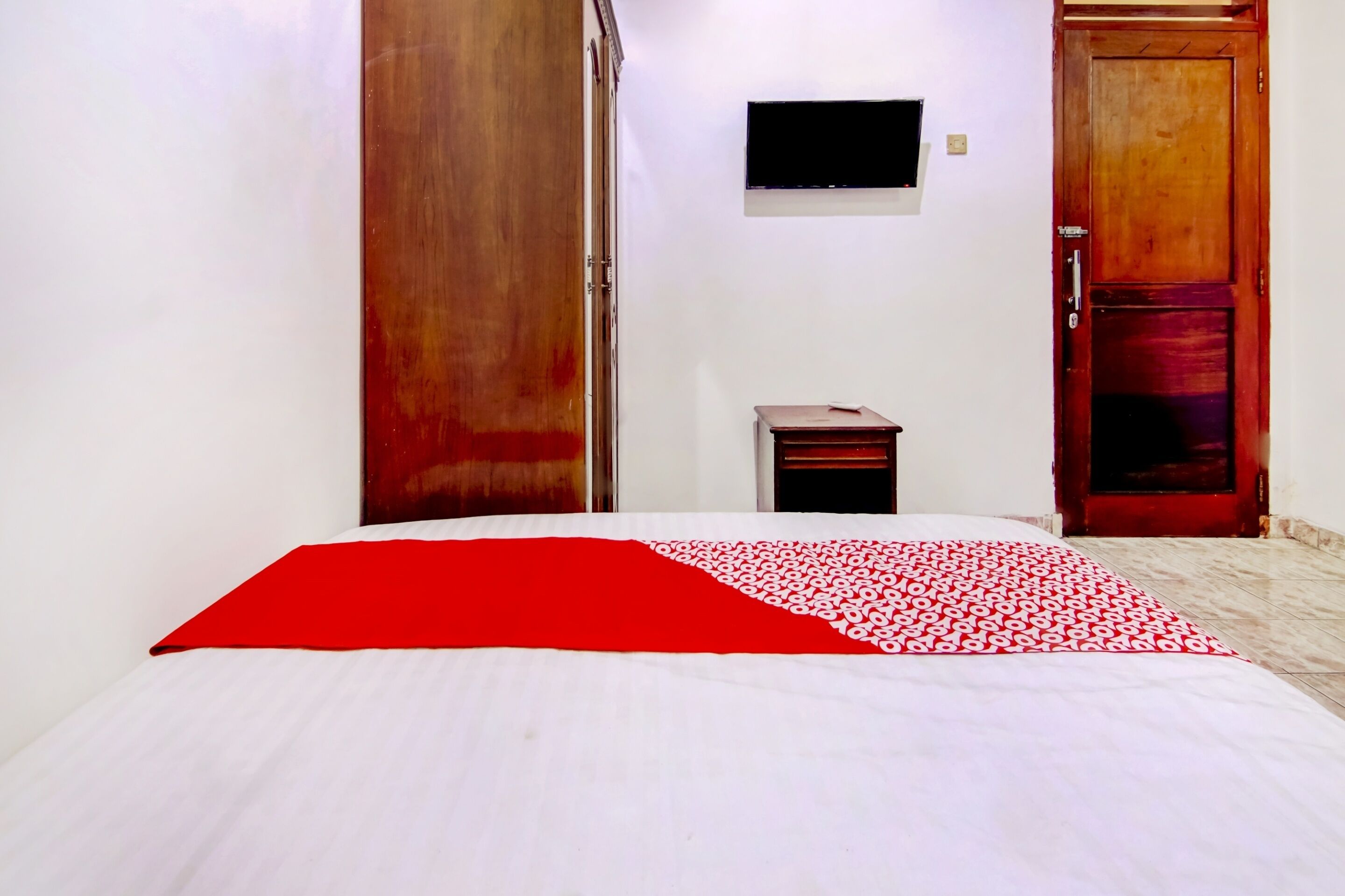 Une chambre d’hôtel avec un lit, une petite table de chevet en bois, un téléviseur fixé au mur et deux portes en bois.