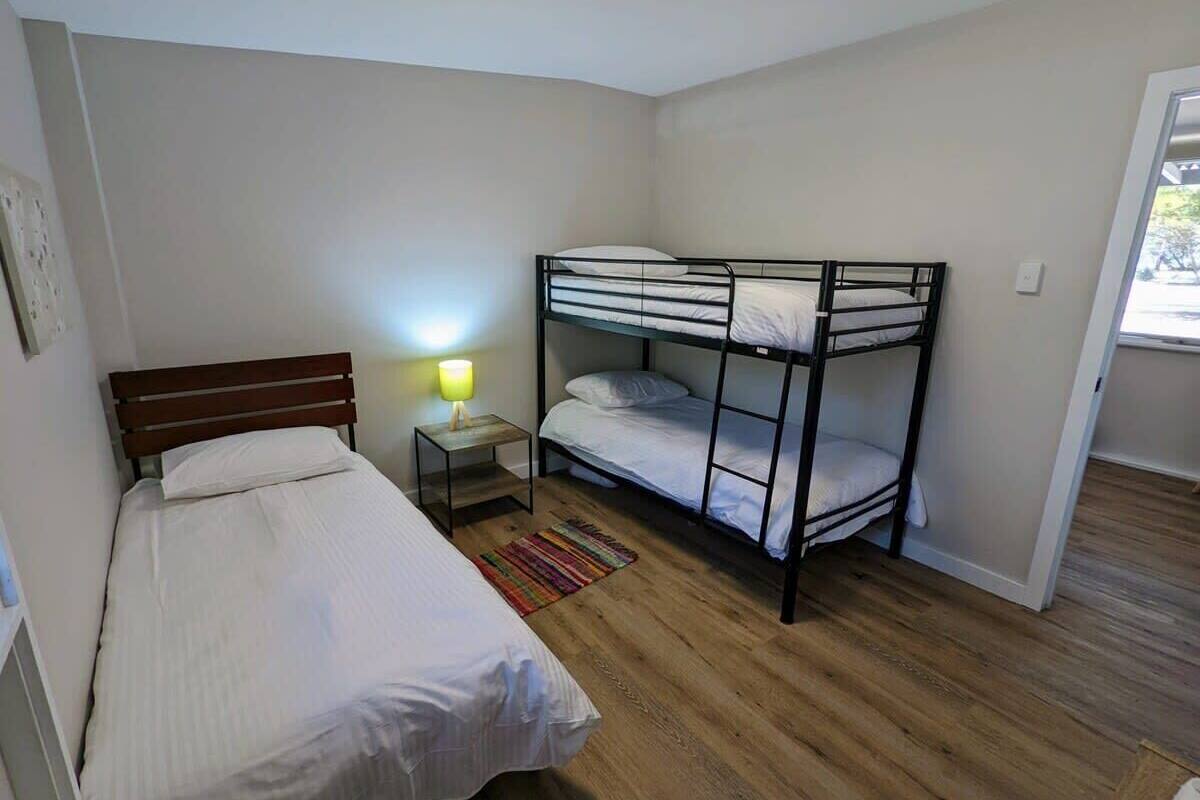 Habitación con dos camas individuales y una litera, suelo de madera, una mesita de noche con lámpara y ventana con vistas.