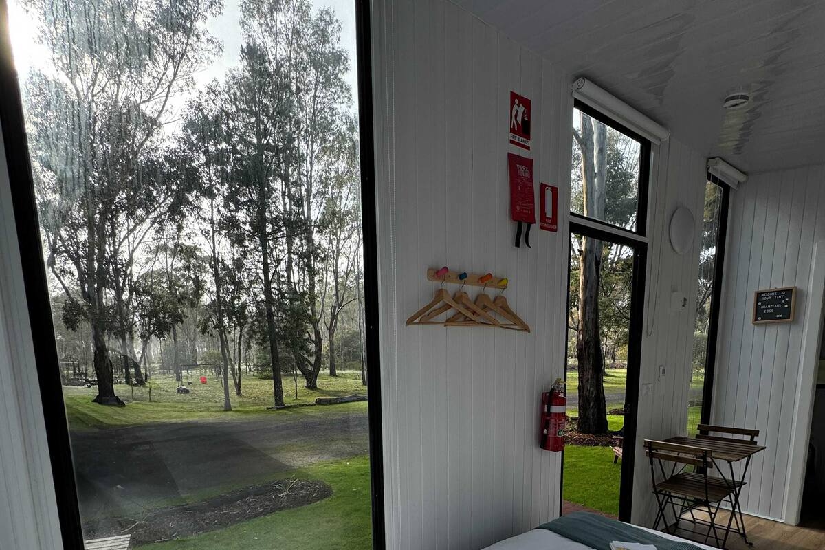 Un dormitorio con un ventanal grande, una cama con ropa blanca, una mesita y un extintor en la pared.