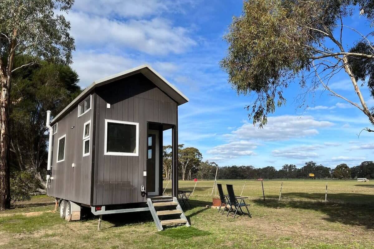 Una cabaña pequeña de un piso con puerta y ventanas, ubicada en un campo cubierto de césped y árboles al fondo.